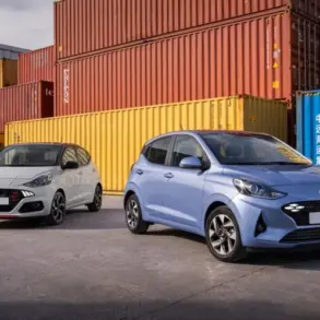 Zwei Hyundai i10 Modelle, einmal in Weiß und einmal in Hellblau, stehen nebeneinander auf einem Industriegelände vor bunten Containern.