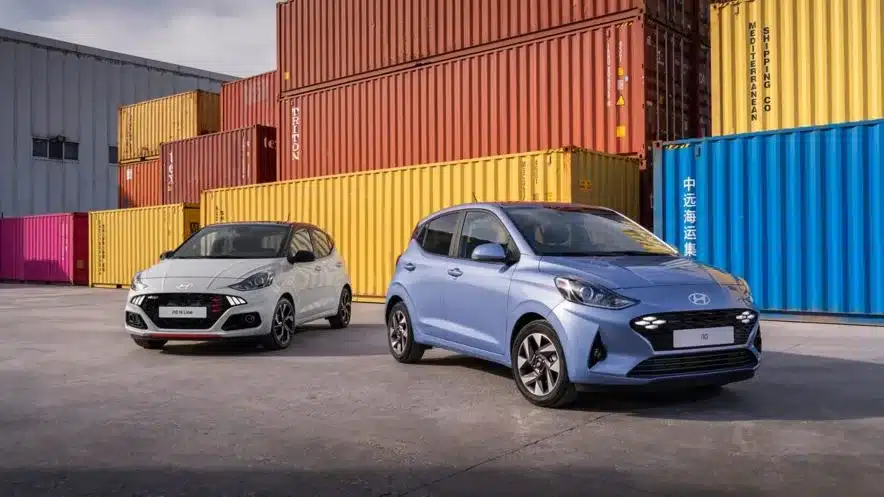 Zwei Hyundai i10 Modelle, einmal in Weiß und einmal in Hellblau, stehen nebeneinander auf einem Industriegelände vor bunten Containern.