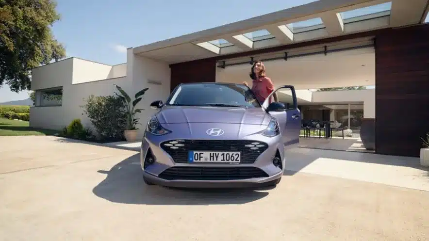 Frau steigt aus einem Hyundai i10 vor einem modernen Haus aus, Frontansicht des Fahrzeugs.