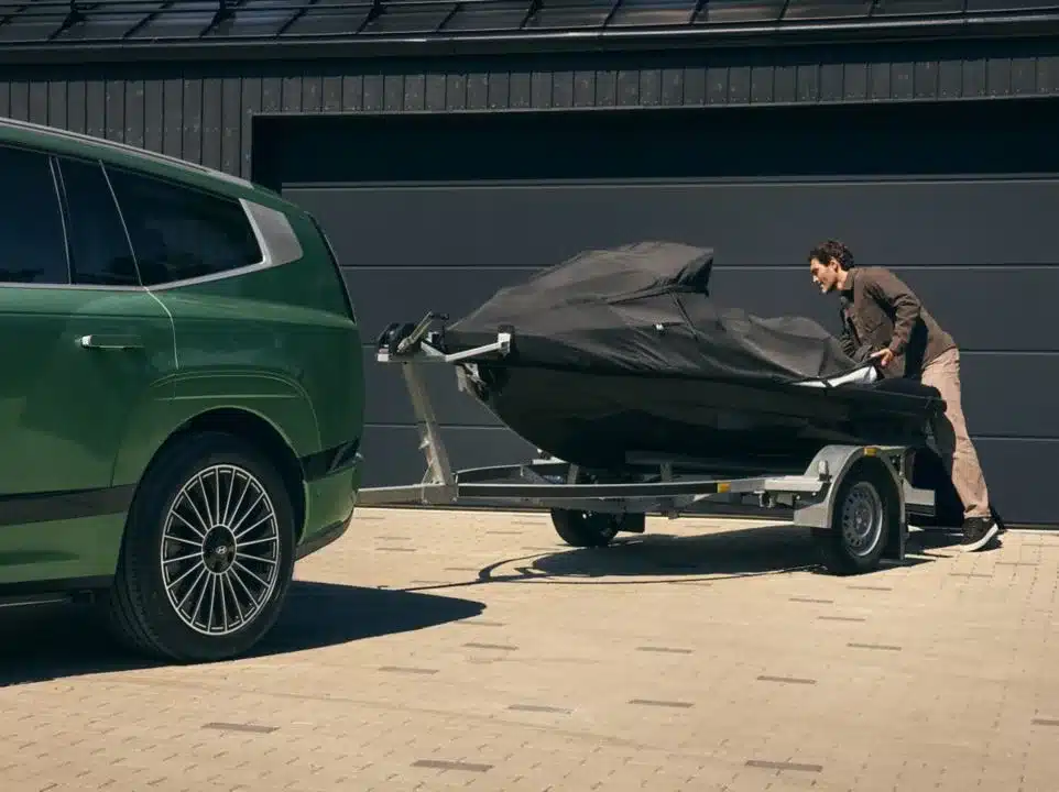 Grüner Hyundai IONIQ 9 zieht einen abgedeckten Jetski auf einem Anhänger vor einer Garage.