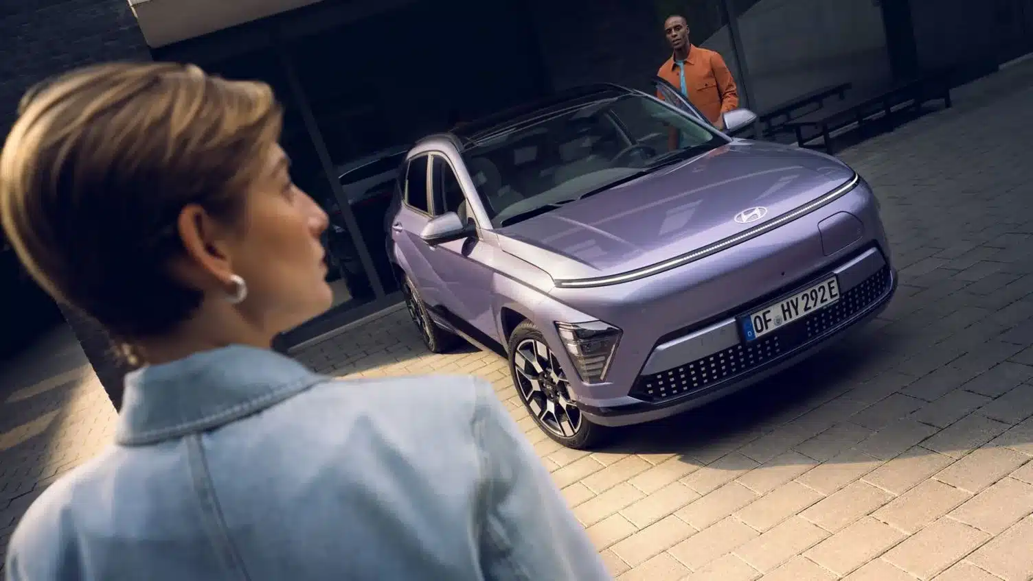 Hyundai KONA Elektro in hellviolett, frontal aufgenommen, mit zwei Personen im urbanen Umfeld.
