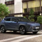 Hyundai KONA in Blaugrau parkt vor einem Stadthotel mit großen Fenstern und Grünpflanzen.
