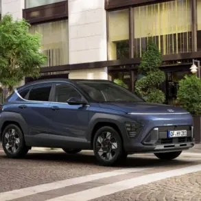 Hyundai KONA in Blaugrau parkt vor einem Stadthotel mit großen Fenstern und Grünpflanzen.
