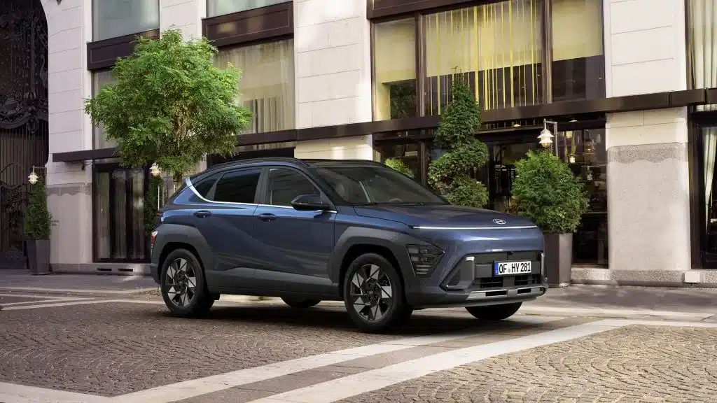Hyundai KONA in Blaugrau parkt vor einem Stadthotel mit großen Fenstern und Grünpflanzen.