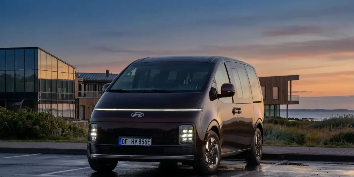 Hyundai STARIA Elektro Van steht auf einem Parkplatz bei Sonnenuntergang vor modernen Gebäuden.