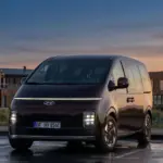 Hyundai STARIA Elektro Van steht auf einem Parkplatz bei Sonnenuntergang vor modernen Gebäuden.