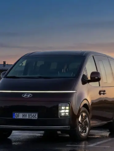 Hyundai STARIA Elektro Van steht auf einem Parkplatz bei Sonnenuntergang vor modernen Gebäuden.