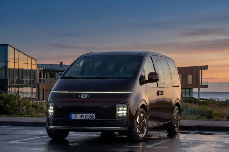 Hyundai STARIA Elektro Van steht auf einem Parkplatz bei Sonnenuntergang vor modernen Gebäuden.