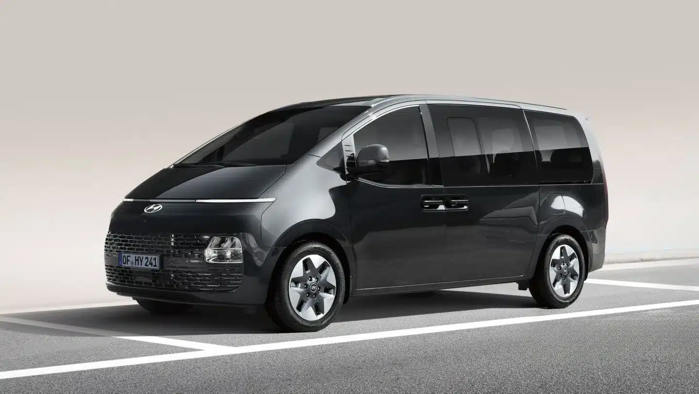 Front- und Seitenansicht des Hyundai STARIA Hybrid Vans in Dunkelgrau auf einer Straße.