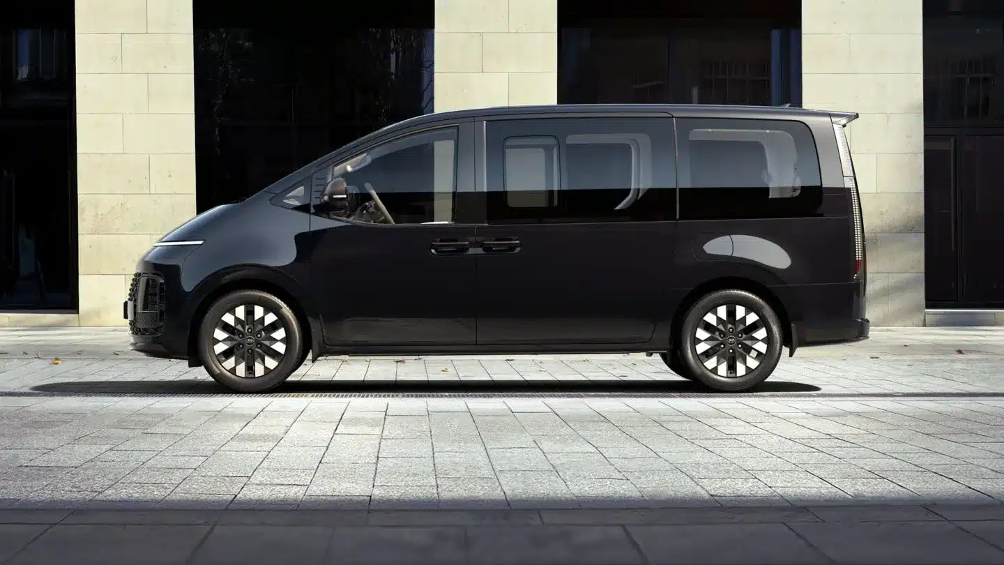 Seitenansicht des Hyundai STARIA Hybrid Vans in dunkler Lackierung vor modernem Gebäude.