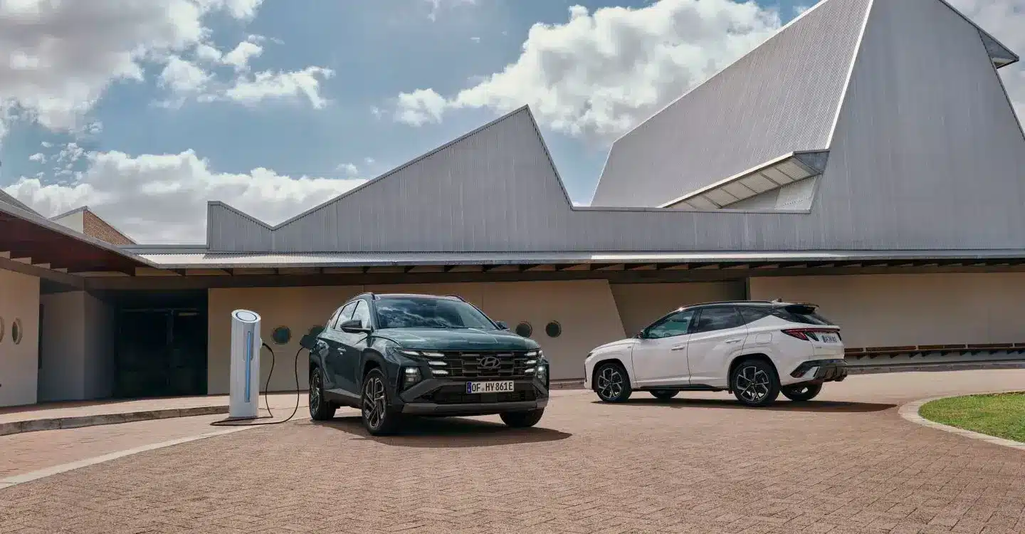 Zwei Hyundai Tucson Plug-in-Hybrid SUV, einer ladend an einer Ladesäule, vor einem modernen Gebäude.