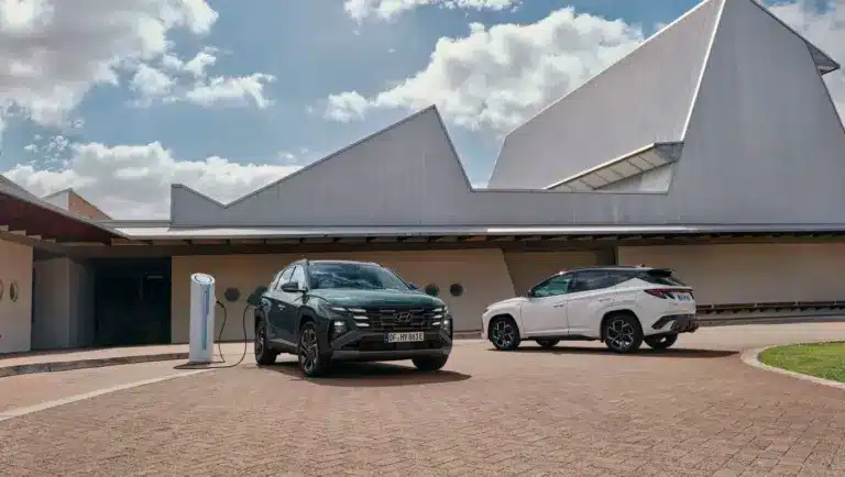 Zwei Hyundai Tucson Plug-in-Hybrid SUV, einer ladend an einer Ladesäule, vor einem modernen Gebäude.