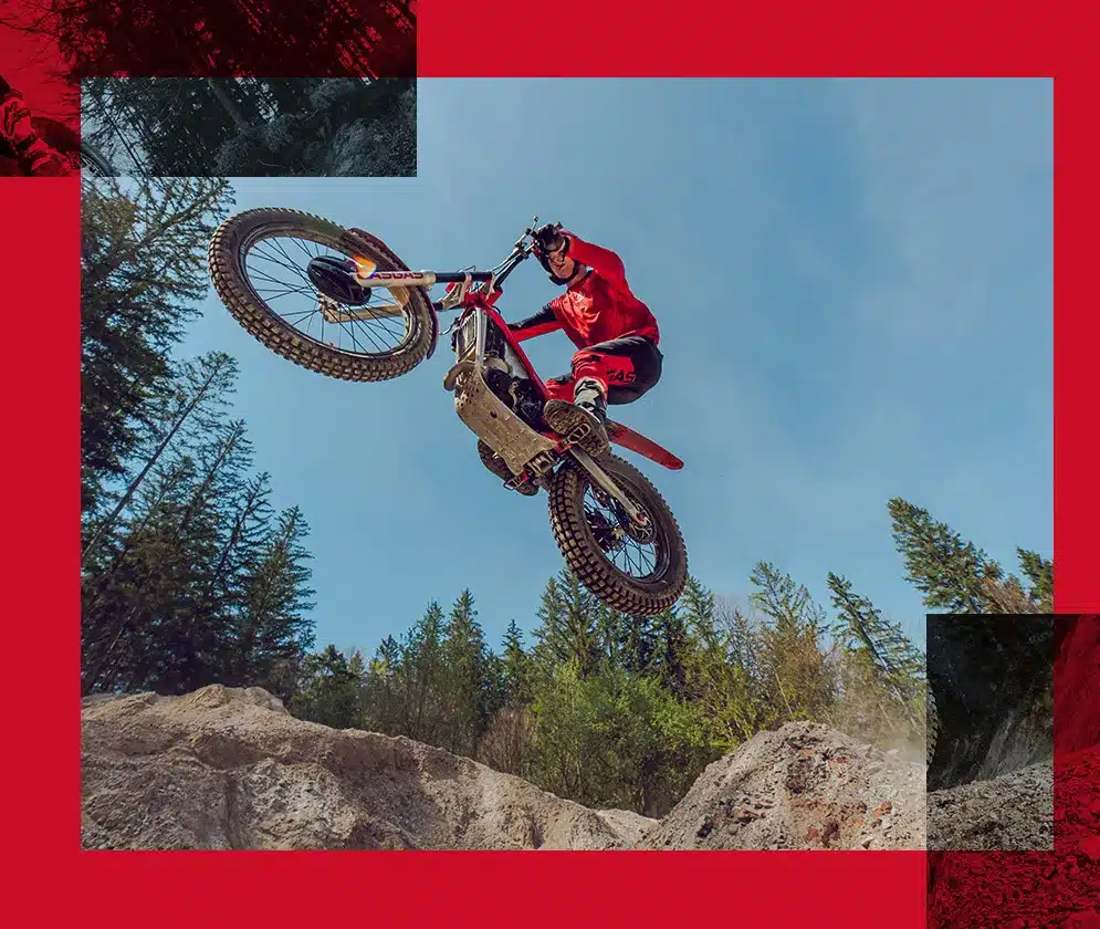 GasGas TXT RACING 125 Trial-Motorrad im Sprung über ein felsiges Gelände, Fahrer in roter Ausrüstung, umgeben von Wald und blauem Himmel.