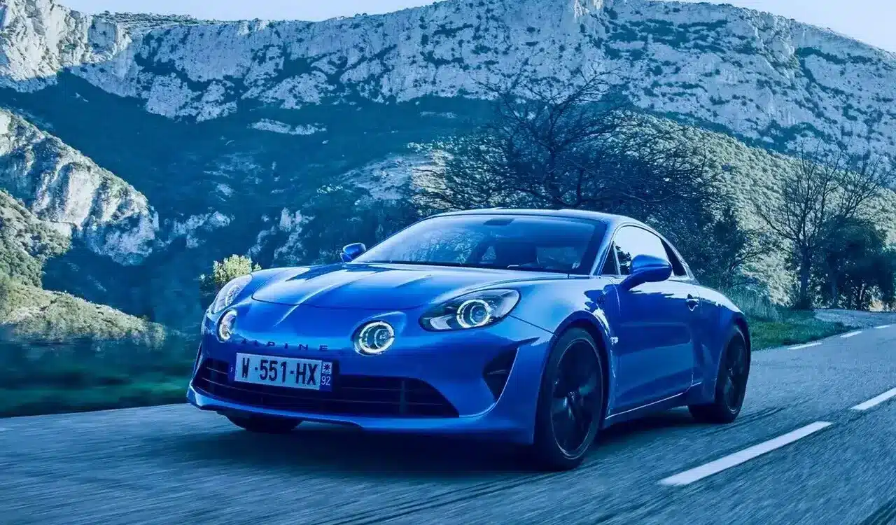 Blauer Alpine A110 fährt auf einer Landstraße vor Bergkulisse.