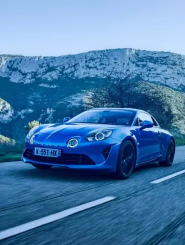 Blauer Alpine A110 fährt auf einer Landstraße vor Bergkulisse.