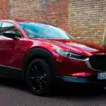 Roter Mazda CX-30 parkt am Straßenrand vor einer Ziegelwand, eine Person geht auf dem Gehweg vorbei
