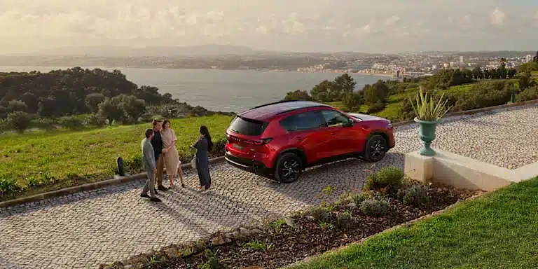 Roter Mazda CX-5 dritte Generation geparkt auf Pflasterweg mit Blick auf eine Stadt und vier Menschen daneben