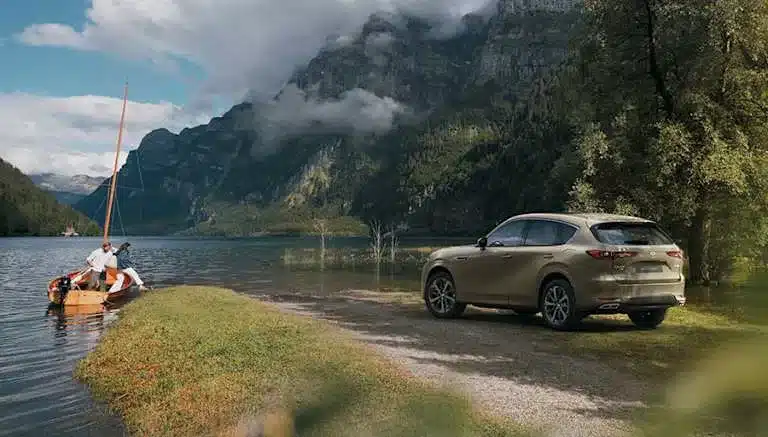 Mazda CX-60 SUV parkt am Wasser mit Bergen im Hintergrund und Segelboot am Steg
