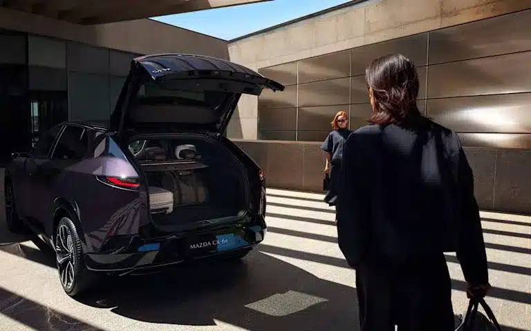 Mazda CX-6e mit geöffnetem Kofferraum, im modernen Stadtdesign, mit Personen im Hintergrund.
