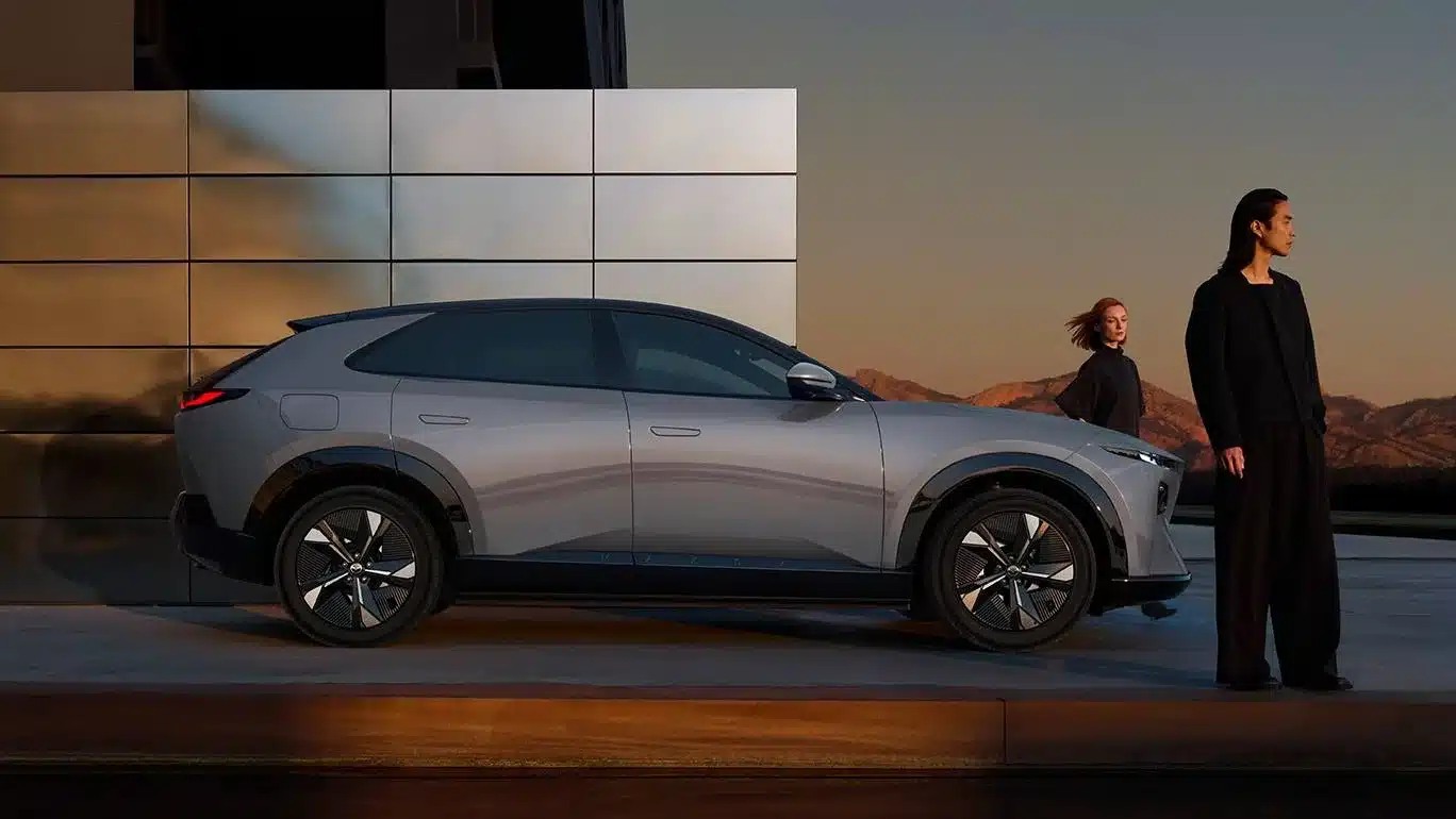 Mazda CX-6e in silbergrau von der Seite mit moderner Fassade im Hintergrund, zwei Personen neben dem Fahrzeug.