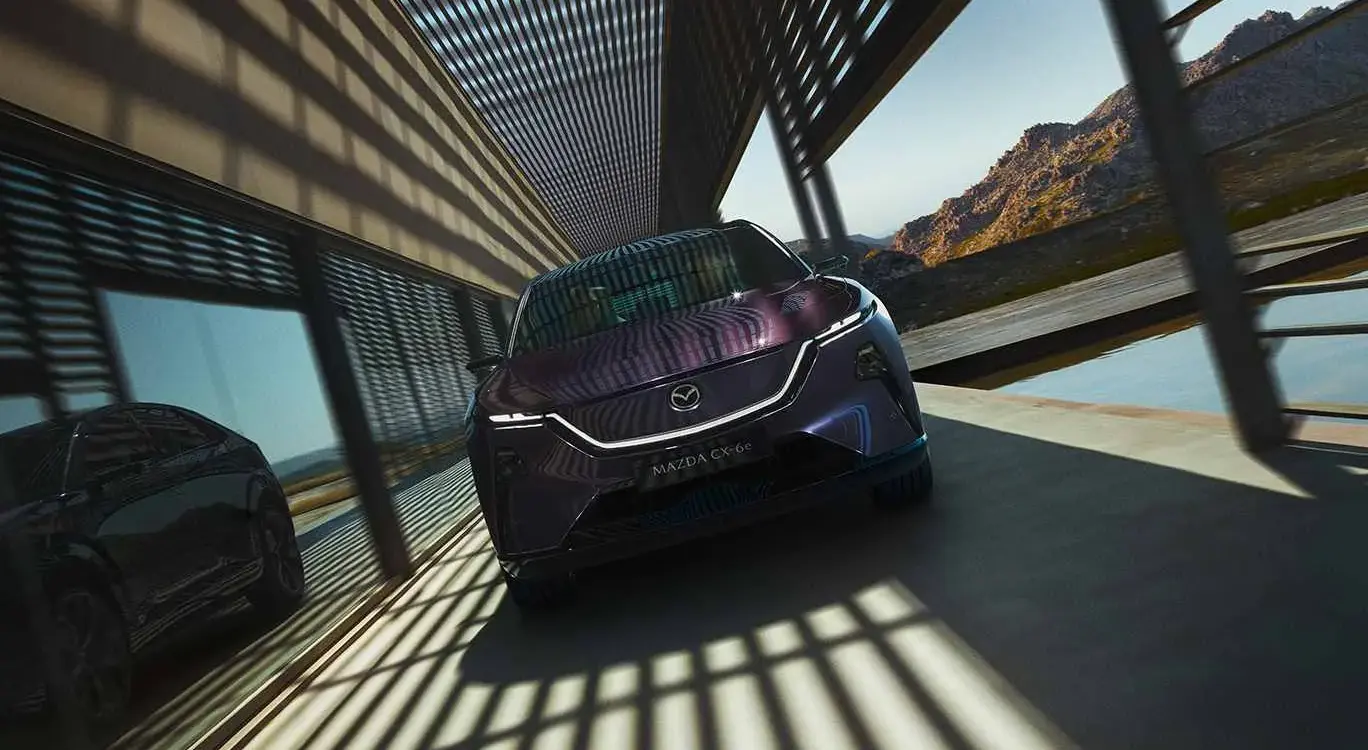 Frontansicht des Mazda CX-6e in violetter Metallic-Lackierung auf einer Terrasse mit Bergen im Hintergrund.