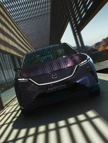 Frontansicht des Mazda CX-6e in violetter Metallic-Lackierung auf einer Terrasse mit Bergen im Hintergrund.