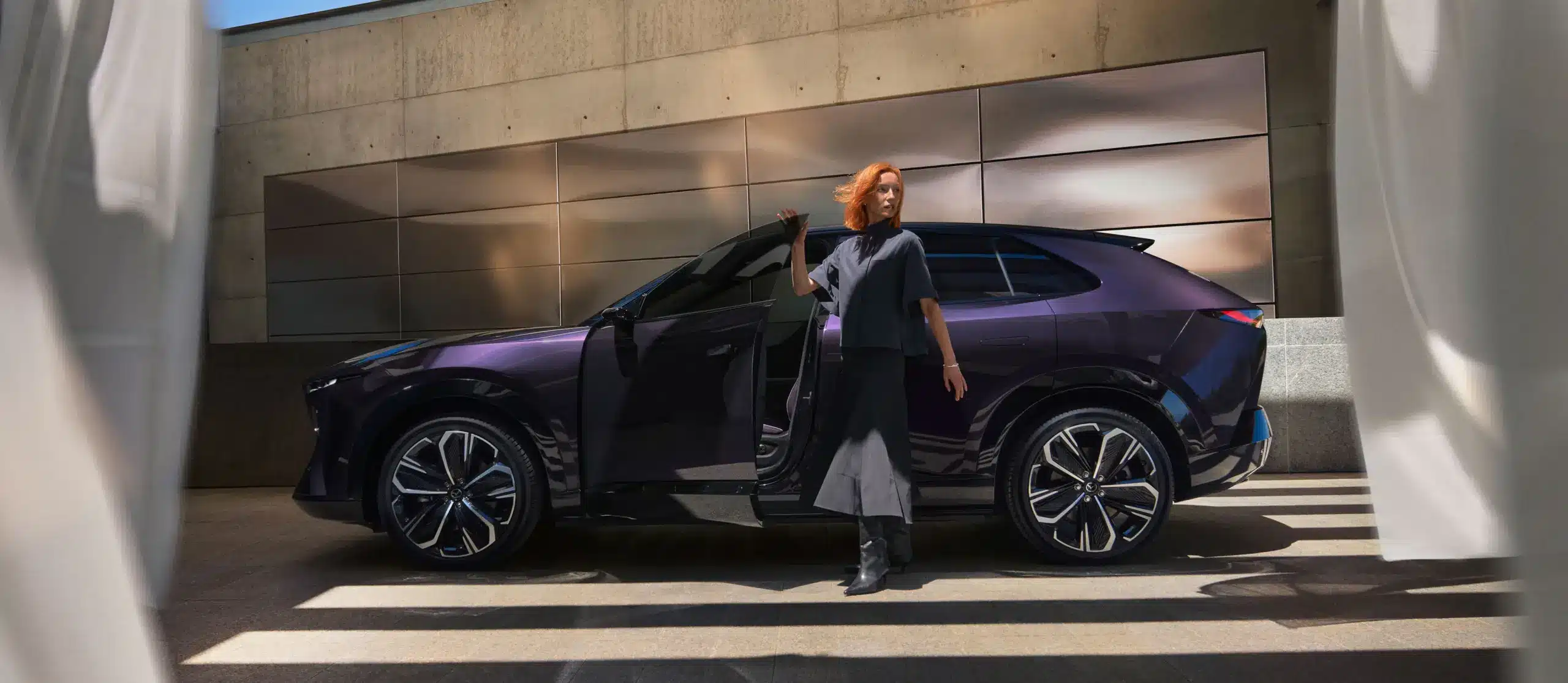 Mazda CX-6e in Nightfall Violet Seitenansicht mit geöffneter Fahrertür, vor urbaner Kulisse.