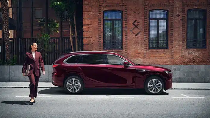 Mazda CX-80 SUV in Rot parkt am Straßenrand vor einer Backsteinfassade, eine Person geht daneben entlang