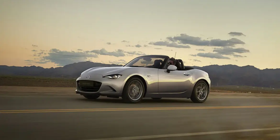 Mazda MX-5 2024 Roadster in Silber fährt bei Sonnenuntergang über eine Landstraße