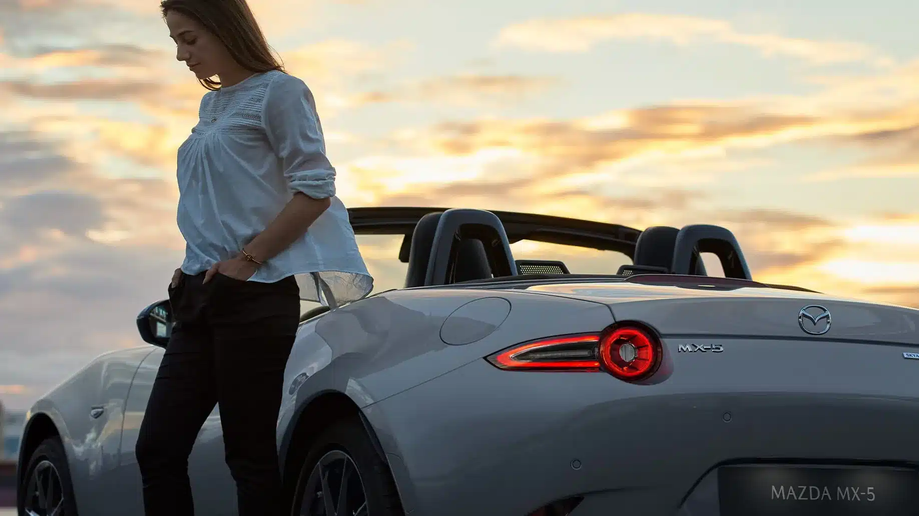 Mazda MX-5 Roadster 2024 in Grau mit offenem Verdeck und einer Frau daneben bei Sonnenuntergang.