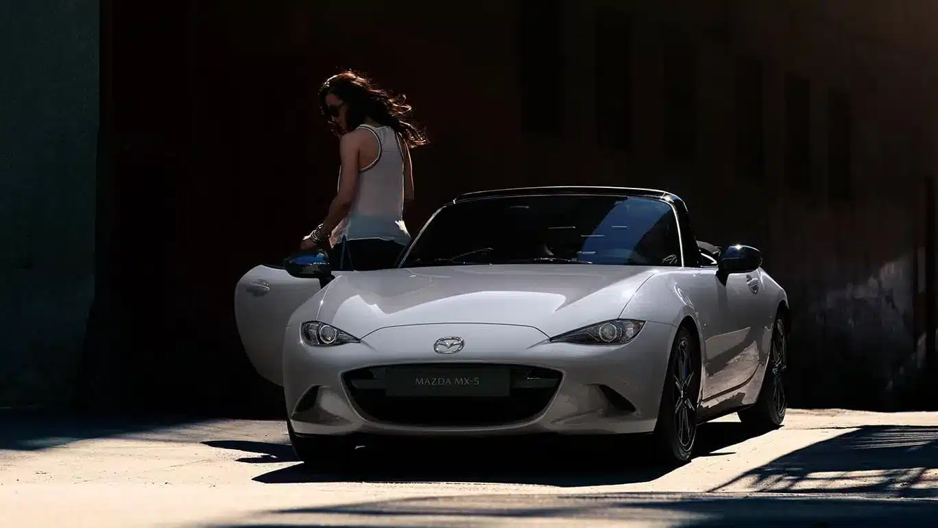 Frontansicht des Mazda MX-5 2024 Roadster in weiß, geparkt mit geöffneter Fahrertür, eine Person steht neben dem Auto.
