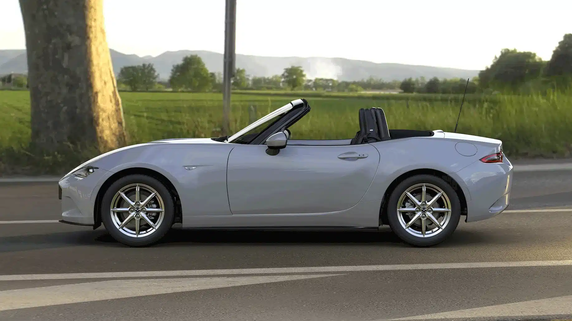 Seitenansicht des Mazda MX-5 2024 Roadster in Aero Grey Metallic mit geöffnetem Stoffverdeck auf einer Landstraße.