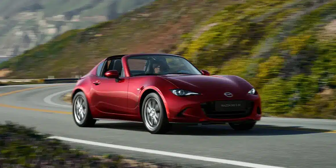 Roter Mazda MX-5 RF fährt auf einer kurvenreichen Straße entlang einer malerischen Landschaft.