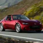 Roter Mazda MX-5 RF fährt auf einer kurvenreichen Straße entlang einer malerischen Landschaft.