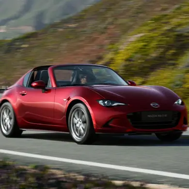 Roter Mazda MX-5 RF fährt auf einer kurvenreichen Straße entlang einer malerischen Landschaft.