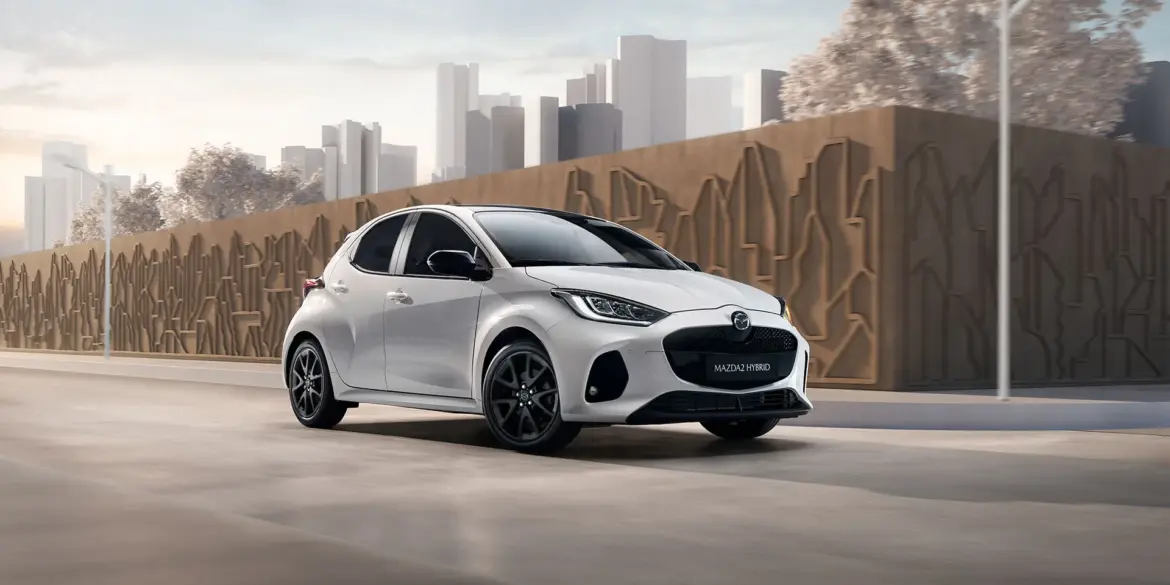 Mazda2 Hybrid 2024 in Weiß fährt auf einer urbanen Straße vor moderner Kulisse.