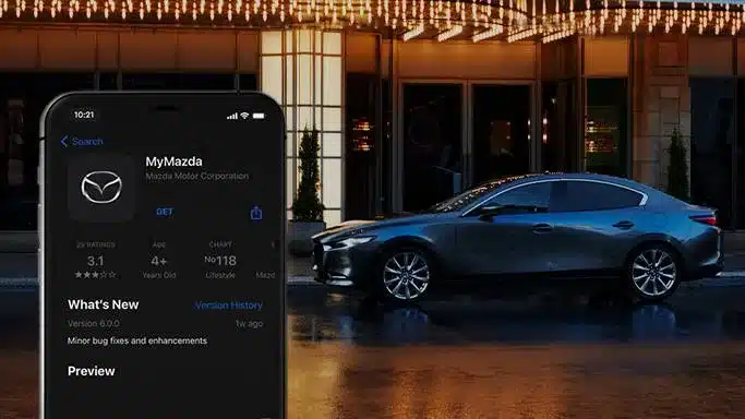 Mazda3 Fastback 2024 in Dunkelgrau seitlich vor einem modernen Gebäude, daneben ein Smartphone mit geöffneter MyMazda App.