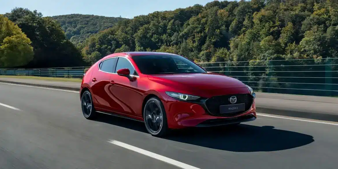 Roter Mazda3 Fünftürer Modelljahr 2024 fährt auf einer Landstraße vor bewaldeter Kulisse.