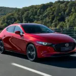 Roter Mazda3 Fünftürer Modelljahr 2024 fährt auf einer Landstraße vor bewaldeter Kulisse.