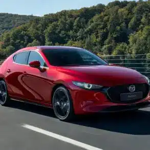 Roter Mazda3 Fünftürer Modelljahr 2024 fährt auf einer Landstraße vor bewaldeter Kulisse.