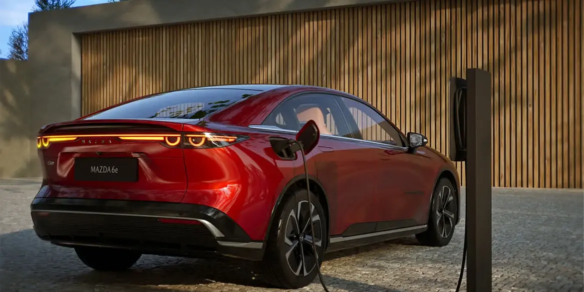 Roter Mazda6e wird an einer Ladesäule geladen, Heckansicht vor Holzfassade.