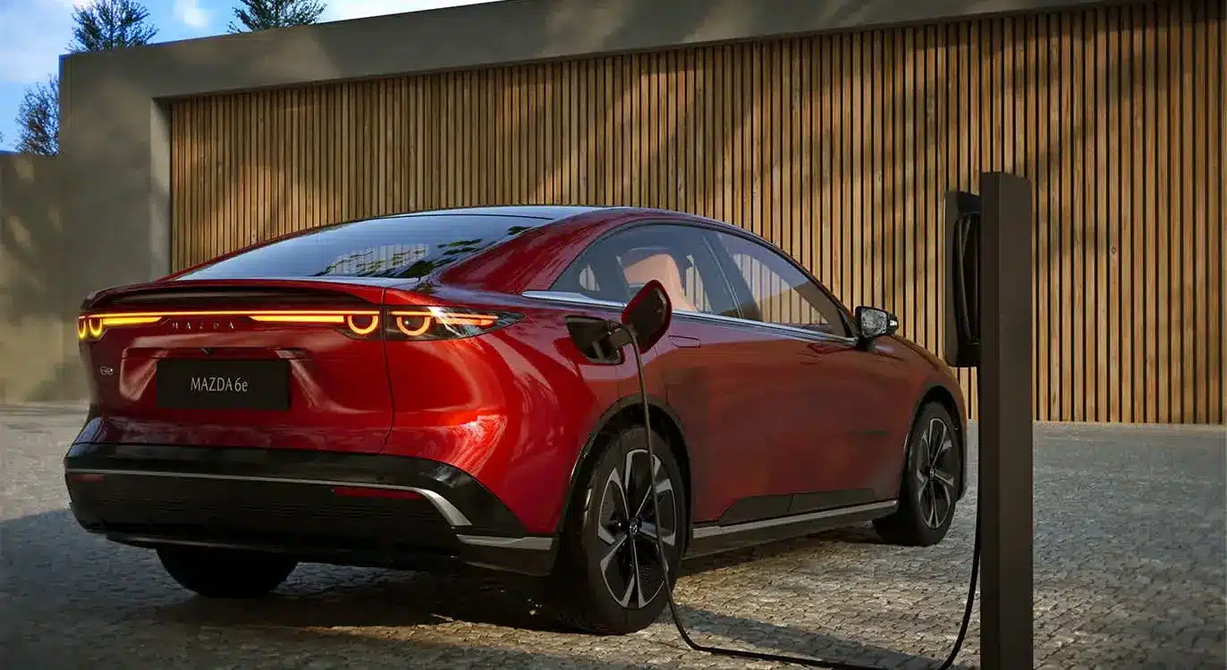Roter Mazda6e wird an einer Ladesäule geladen, Heckansicht vor Holzfassade.