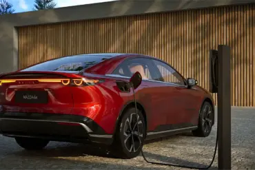 Roter Mazda6e wird an einer Ladesäule geladen, Heckansicht vor Holzfassade.