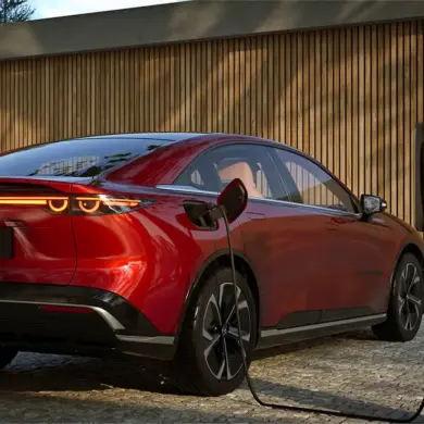 Roter Mazda6e wird an einer Ladesäule geladen, Heckansicht vor Holzfassade.