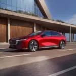 Rote Mazda6e vollelektrische Limousine fährt auf einer modernen Straße vor einem Gebäude.