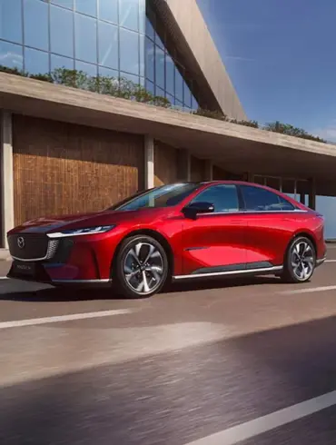 Rote Mazda6e vollelektrische Limousine fährt auf einer modernen Straße vor einem Gebäude.