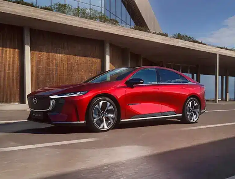 Rote Mazda6e vollelektrische Limousine fährt auf einer modernen Straße vor einem Gebäude.