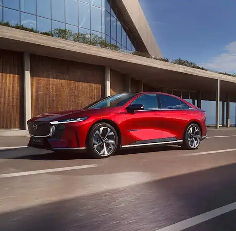 Rote Mazda6e vollelektrische Limousine fährt auf einer modernen Straße vor einem Gebäude.