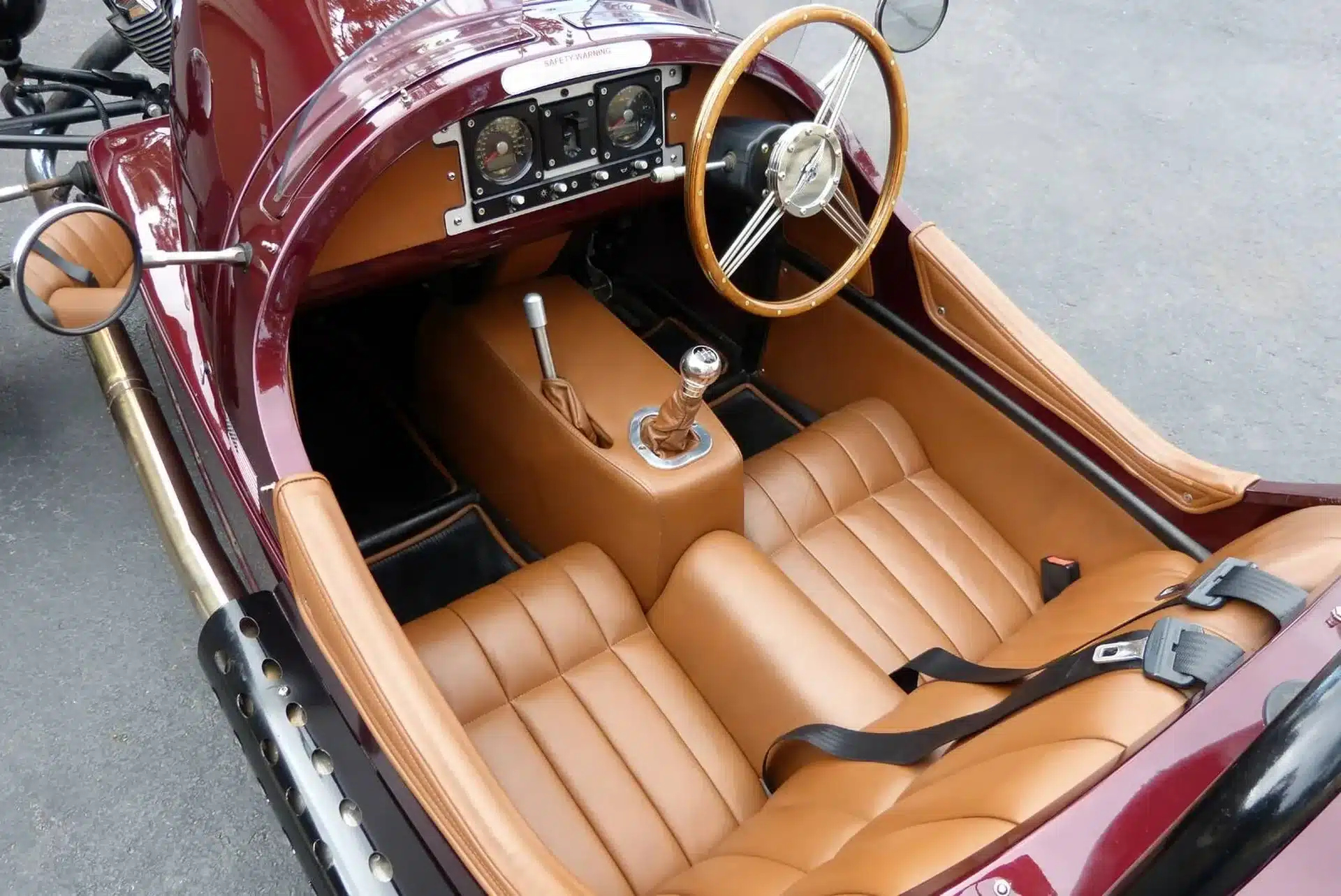 Innenraum eines Morgan Three-Wheeler mit beigen Ledersitzen, Holzlenkrad und zentraler Handschaltung.