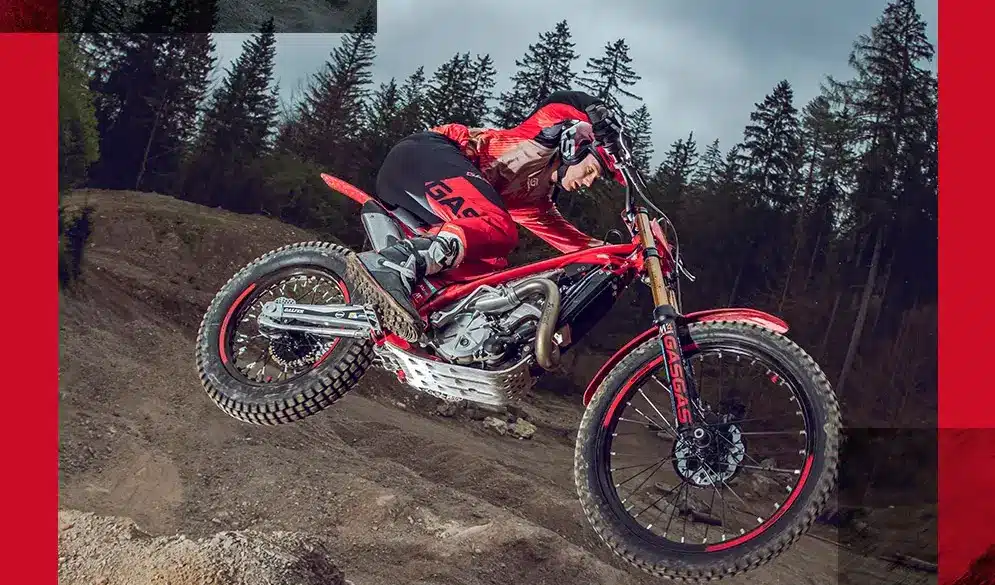 GASGAS TXT GP 250 2025 Trial-Motorrad im Sprung im Gelände, Fahrer mit roter Ausrüstung, Wald im Hintergrund.